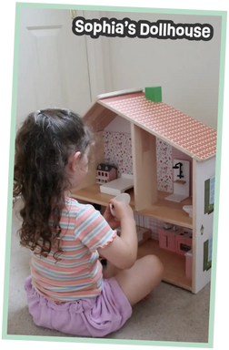 A wooden dollhouse with a pink tiled roof, featuring multiple rooms with furniture including a bed, sink, and kitchen cabinets.